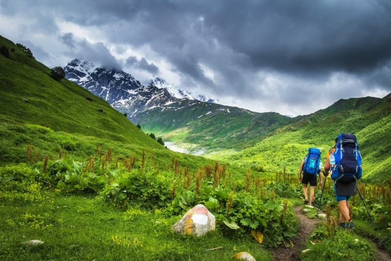 Tourist with backpack hike on mountain trek. Hiking in Svaneti, Georgia. Trekking in mountains. Two men walk on grassy hills. Leisure activity. Sport tourism. Adventures of hikers. Climbers in mounts